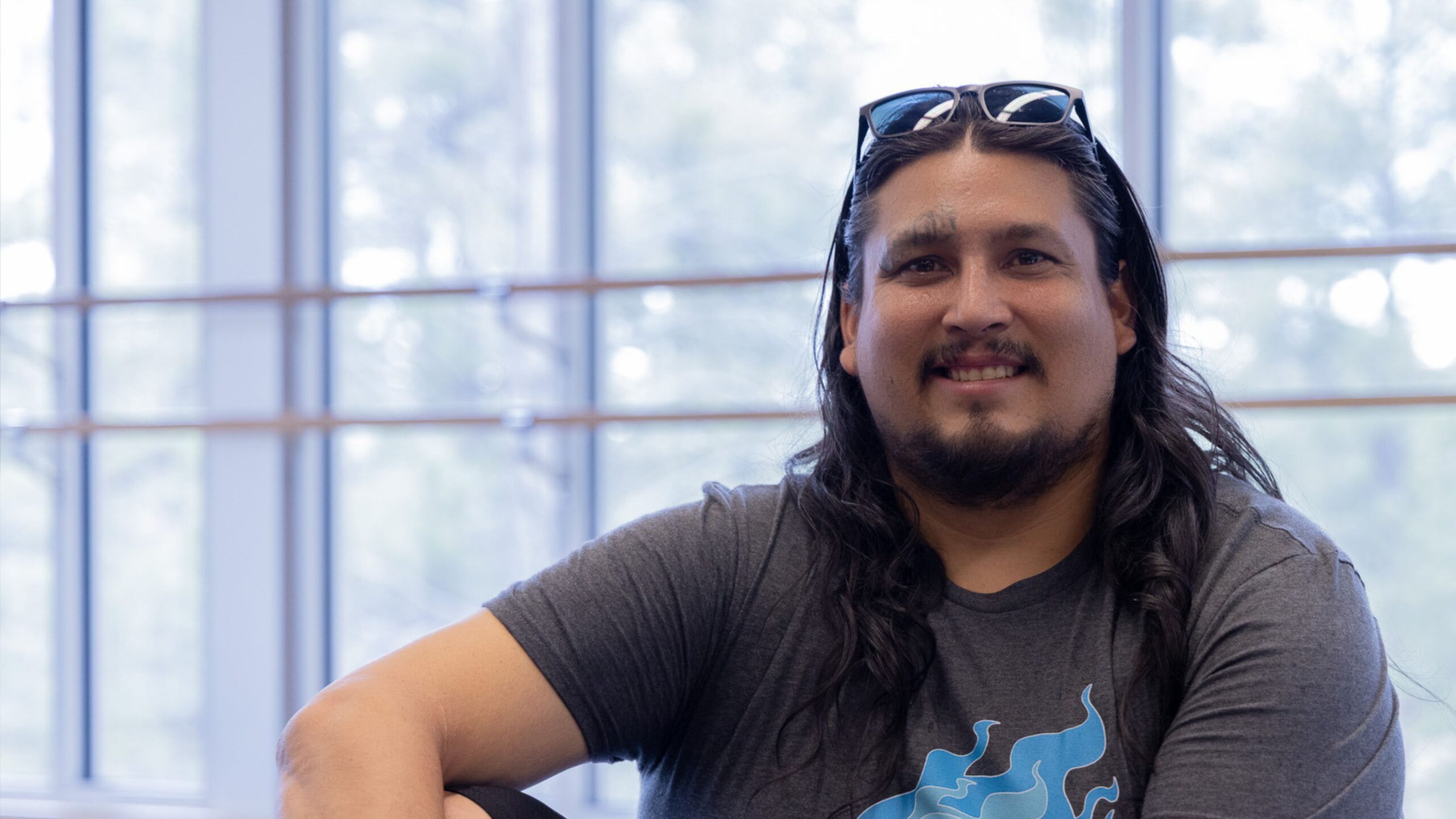 A CCC student smiles while seated in a bright campus building, with large windows and trees visible in the background.